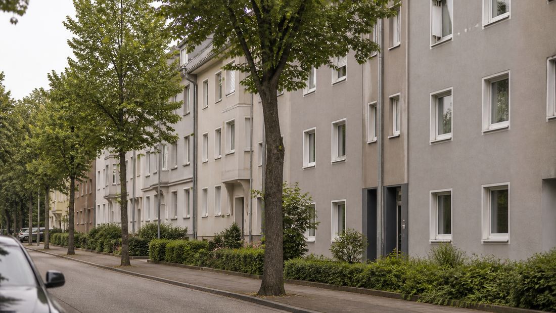 Wohnstraße mit einer Zeile dreigeschossiger Wohnhäuser, Straßenbäumen und Heckenstreifen